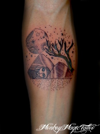 Tatuaje de un paisaje estrellado, con un árbol, la luna montañas y una cara grabada en ellas