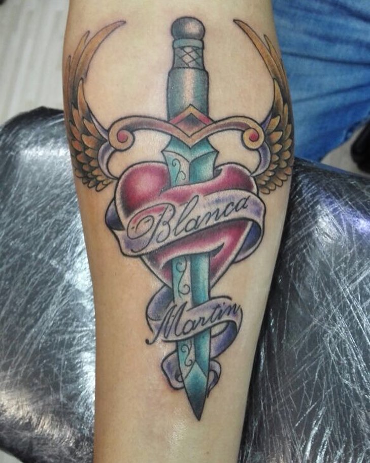 Tattoo de un puñal con alas atravesando un corazón y una cinta con nombres
