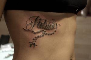 Tatuaje de un nombre con un rosario colgando en las costillas de una mujer