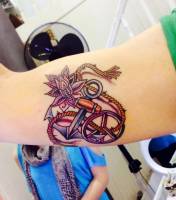 Tatuaje a color en el brazo de un ancla junto al logo hippie y una flor